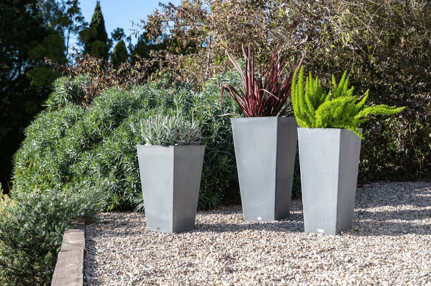 Three tall tapered square planters in a garden, showcasing durable recycled materials and vibrant plants.