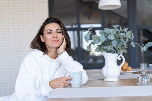 Young woman in a white hoodie enjoying a cup of tea at a table with greenery and pastries.