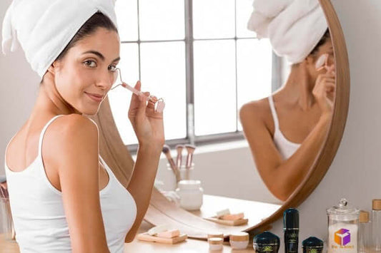 Woman with a towel on her head applying skincare in front of a mirror, surrounded by beauty products.