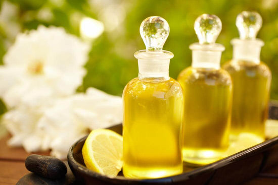 Three glass bottles of golden body oil with lemon slices, set against a blurred greenery background.