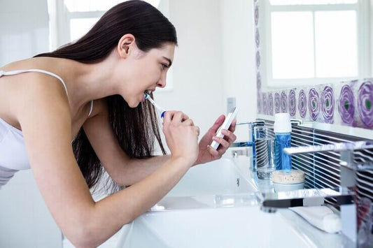 Woman brushing her teeth while checking her phone in a modern bathroom setting.