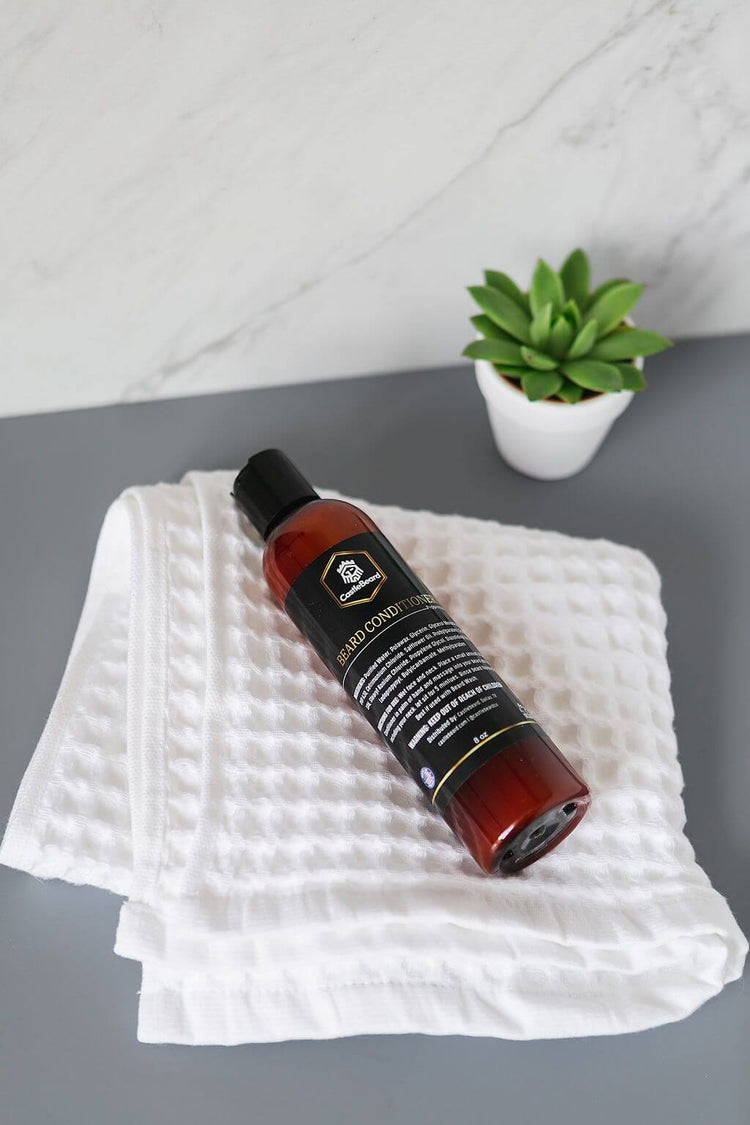 Beard conditioner bottle on a textured white towel with a small green plant in the background.