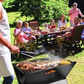 Portable BBQ grill with delicious food being prepared outdoors, surrounded by people enjoying a cheerful gathering.