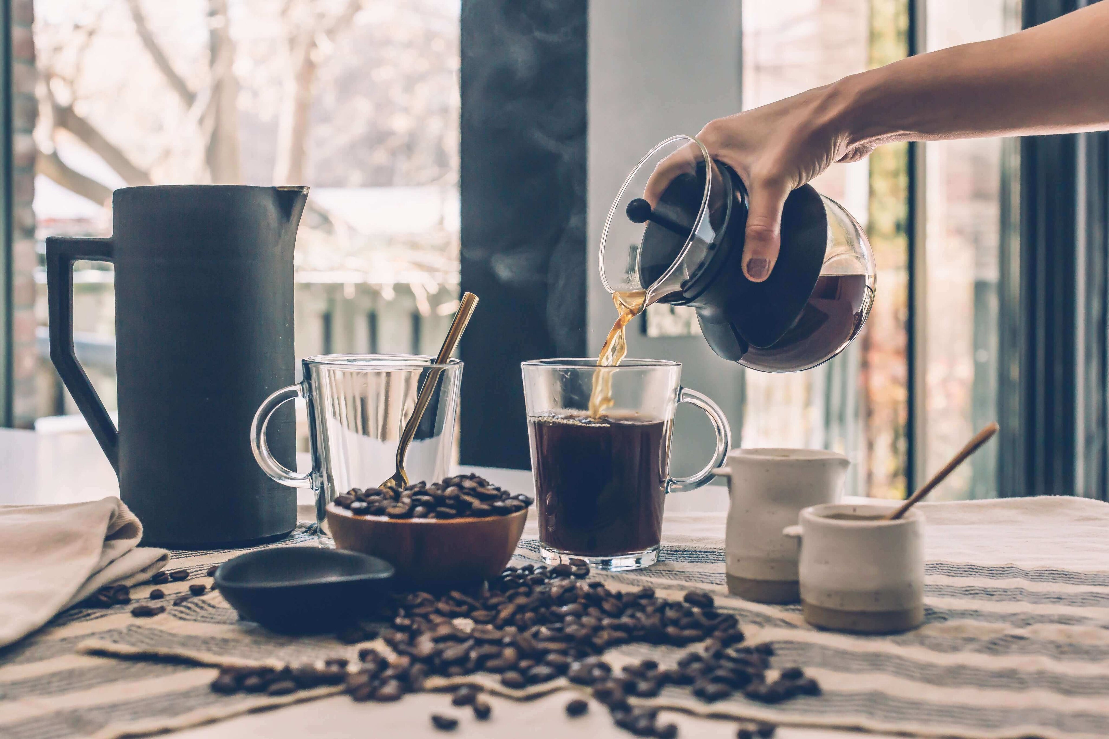 A person pouring freshly brewed coffee into a glass cup, with coffee beans and brewing tools on a table.