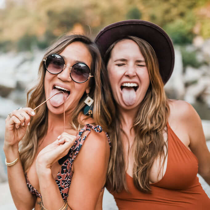 Two women smiling and sticking out their tongues, showcasing fun moments outdoors with sunglasses and summer outfits.