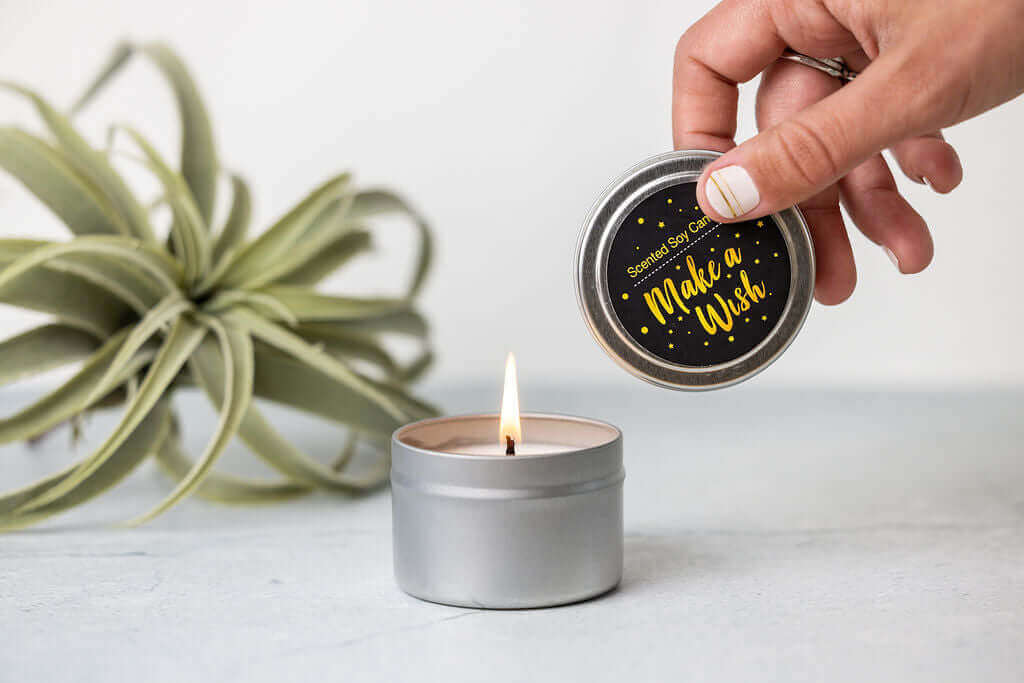 Person opening a silver candle tin labeled 'Make a Wish' with a lit candle inside, surrounded by a succulent plant.