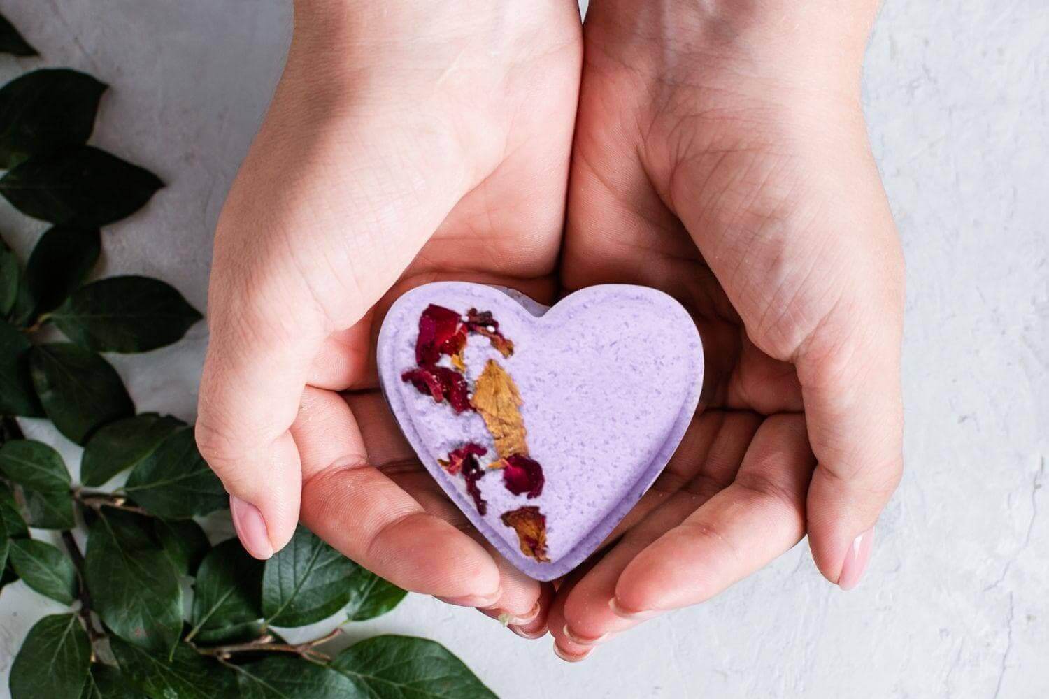 Hands holding a heart-shaped lavender shower steamer with dried petals, surrounded by greenery.