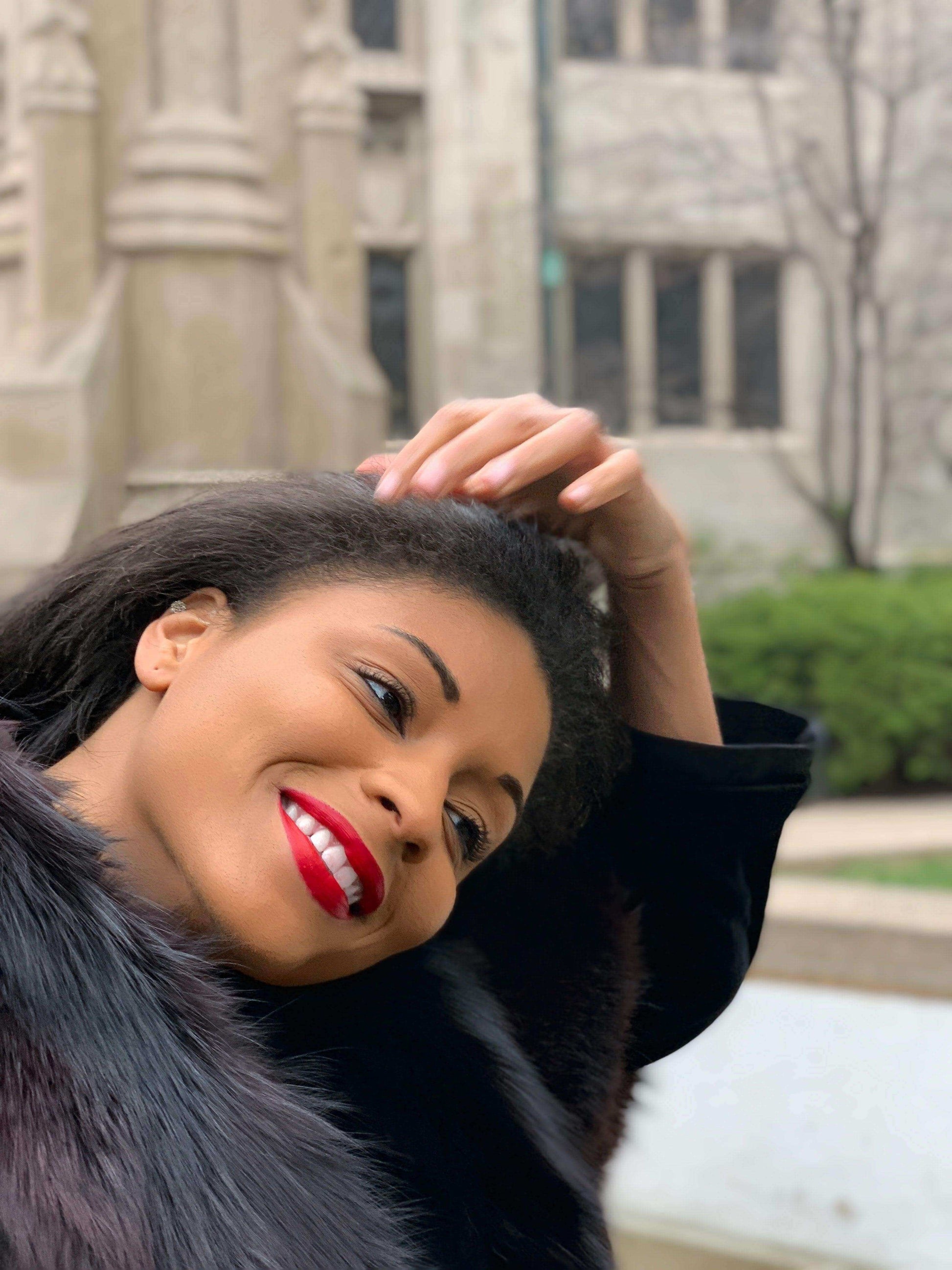 Smiling woman with red lips touching her hair, wearing a black coat near lush greenery.