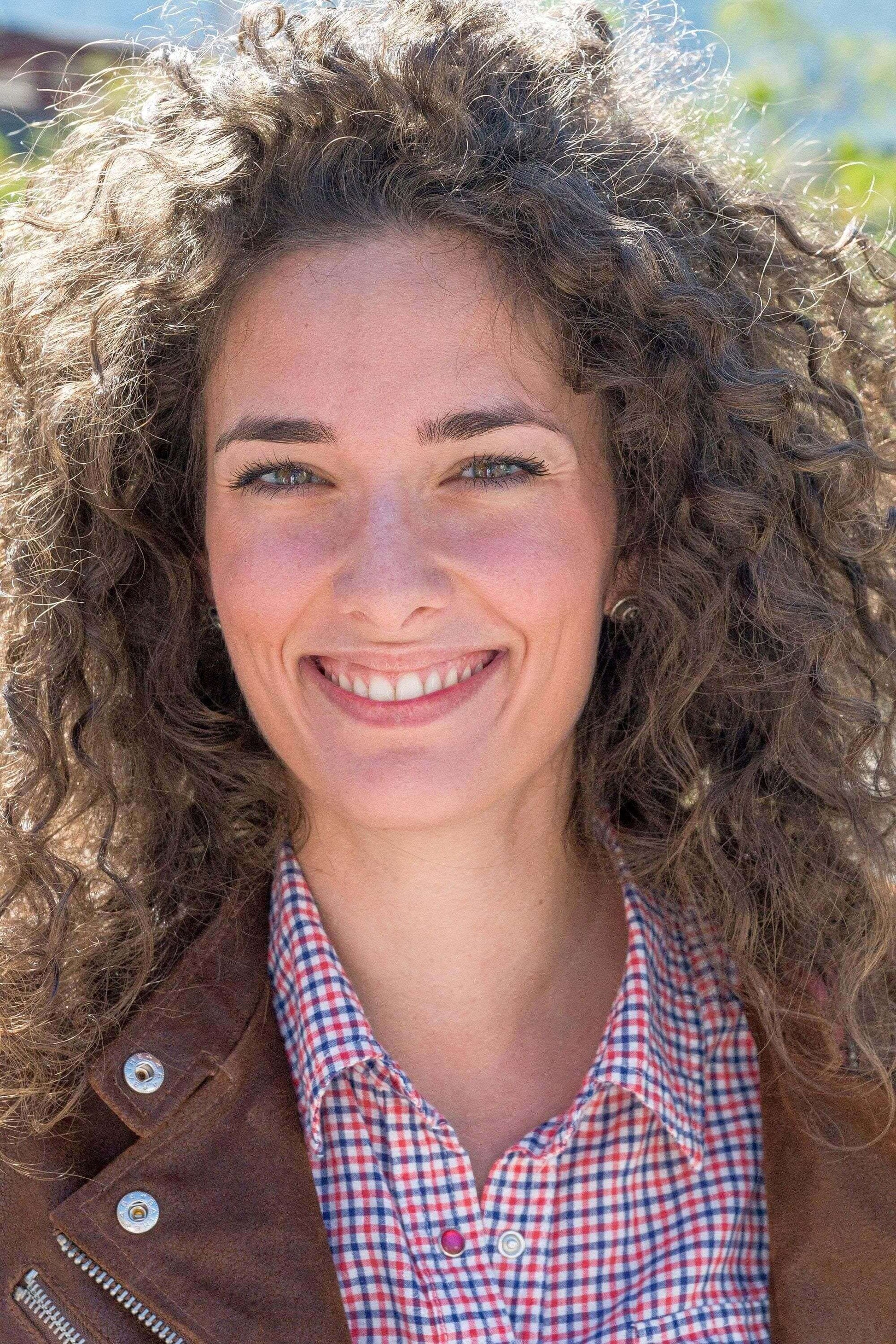 Smiling woman with curly hair wearing a checkered shirt and brown jacket outdoors.
