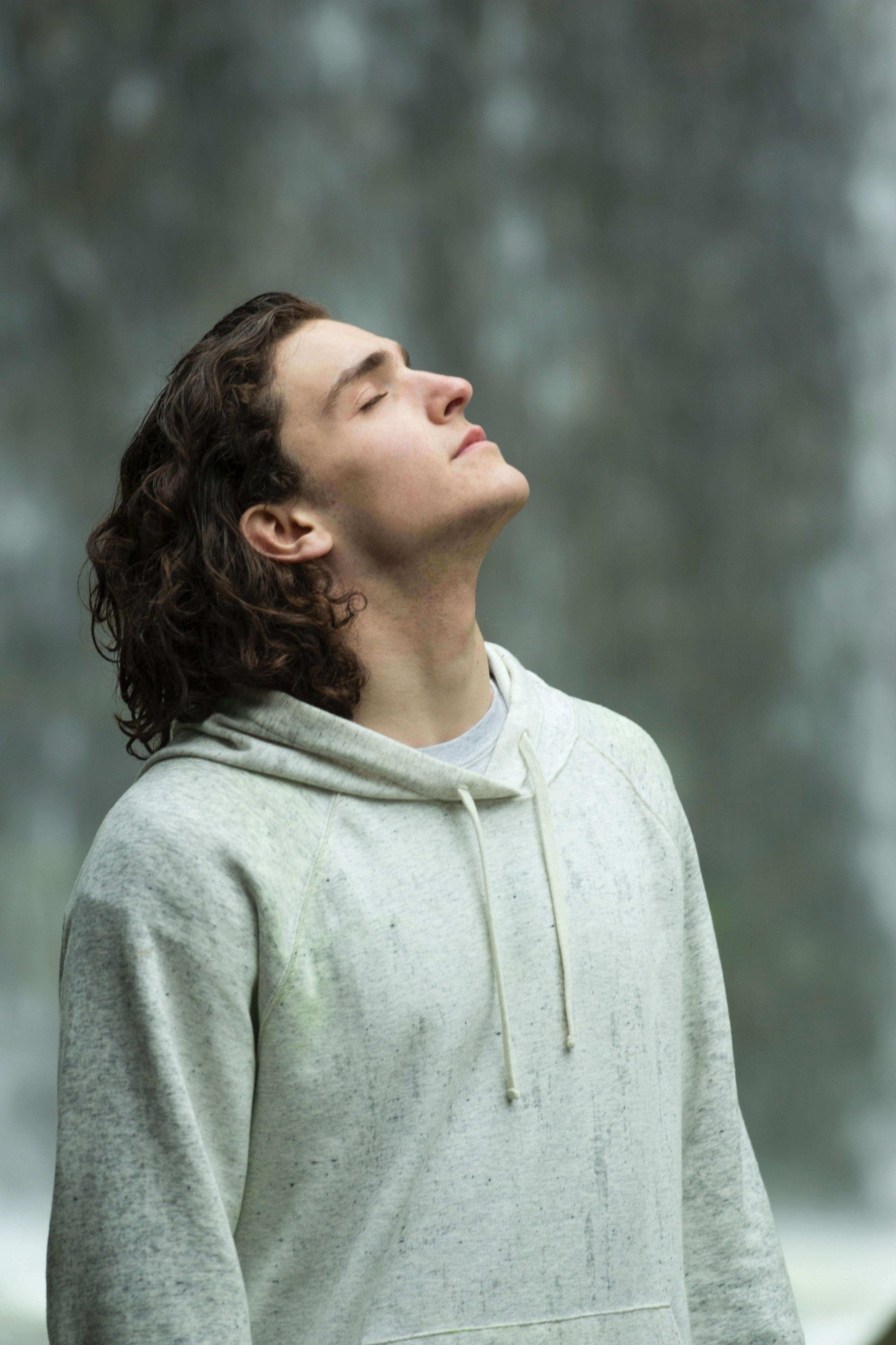 Man with curly hair wearing a gray hoodie standing in front of a beautiful waterfall, eyes closed in tranquility.