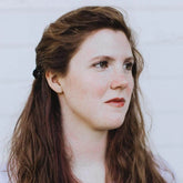 Close-up of a woman with long brown hair and soft makeup, gazing thoughtfully against a light background.